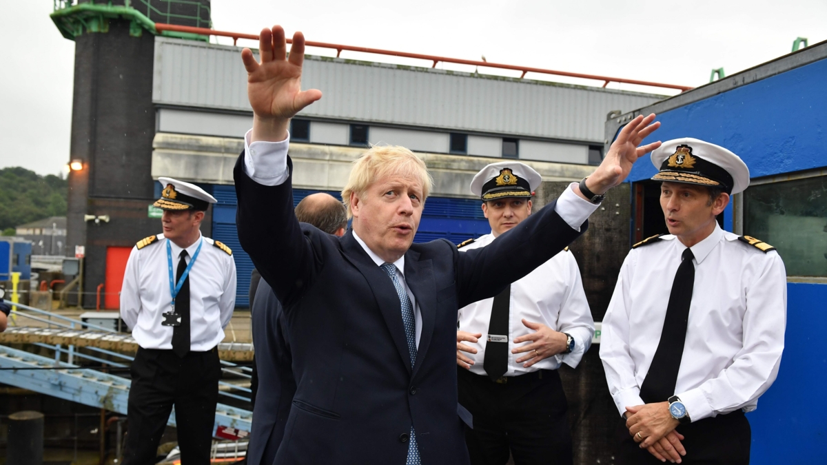 Bildet viser Storbtiannias statsminister Boris Johnson i halvfigur som gestikulerer med hendene i været. I bakgrunnen fire menn i marineuniformer.