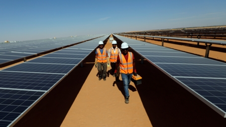 Bildet viser tre arbeidere som går mellom solcellepanelene på solkraftstasjonen Benban i Aswan, Egypt.